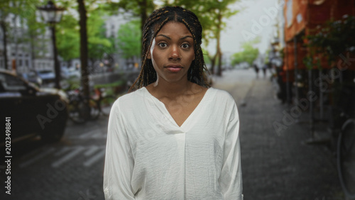 Bild auf Leinwand Woman shrugs shoulders on street in white blouse, braided hair framing face, slight grimace; uncertain hesitation