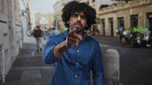 Fotografía Man points finger directly at camera on busy city street near bollard and parked motorcycles;accusation anger confrontation