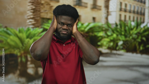Canvas Print Man covering ears with hands in front of building courtyard wearing red polo shirt and frowning in sunlight; sensory overload frustration