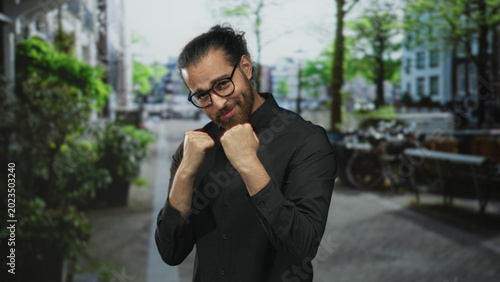 Photos Man with fists raised on street wearing black shirt and glasses, punching toward camera; confidence fitness training