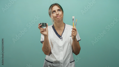 Bild auf Leinwand Woman holding a cream jar and paintbrushes, wearing white uniform and raising hands while looking up in studio; thoughtful decision