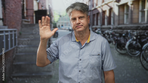 Foto Man raising hand in a casual wave on a narrow urban street with bicycles and brick steps visible; friendly greeting