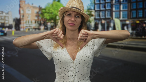 Quadro su tela Woman showing a thumbs down gesture on a city street wearing a straw hat and polka dot dress with visible cleavage and neutral expression; disapproval