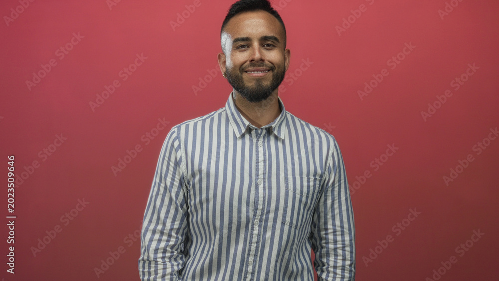 Obraz premium Man waving right hand in studio with red backdrop and blue white striped shirt, smiling at camera; friendly greeting.