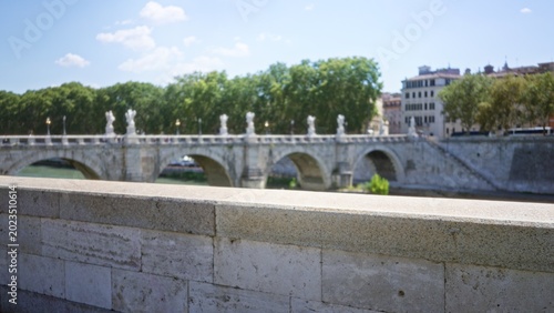 Billede på lærred Stone parapet in sharp focus with historic roman bridge and riverside trees softly blurred in shallow bokeh street; background backplate copyspace calm