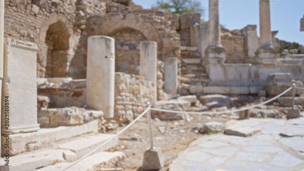 Fototapeta premium Ancient stone ruins and weathered marble columns lining a sunlit path, softly blurred outdoor background; background backdrop copyspace calm.