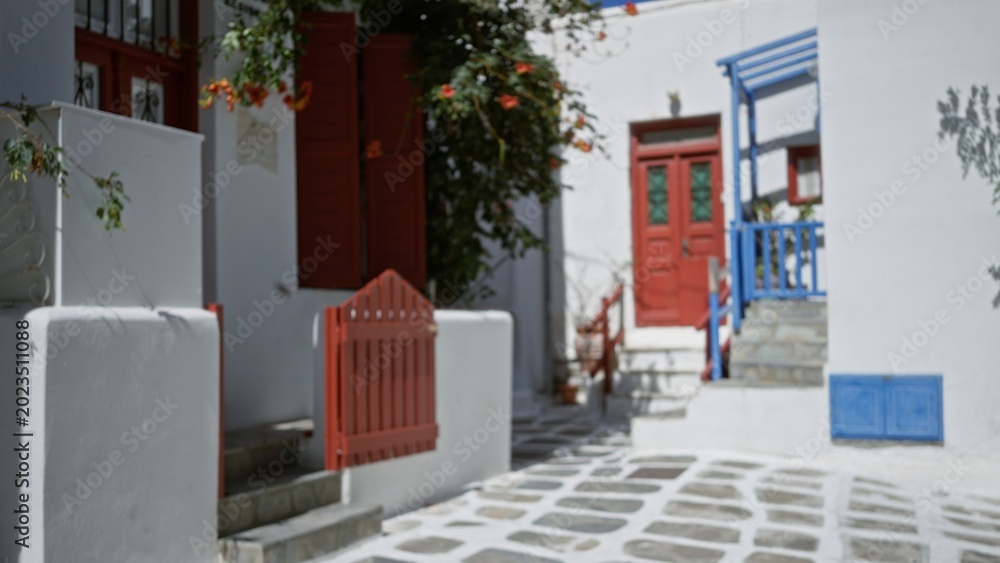Fototapeta premium Sunlit mediterranean alley with whitewashed walls, red doors and stone paving, softly blurred bokeh; background backdrop copyspace calm.