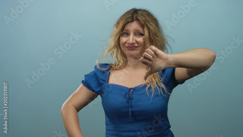 Obraz na plátně Woman showing thumbs down gesture in studio with teal backdrop and wearing blue dress with tousled blonde hair; disapproval