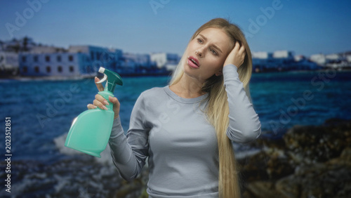 Foto Woman holding spray bottle, hand to head and eyes closed, long blonde hair, standing in front of coastal building by the sea with rocky shore; annoyance cleaning hesitation