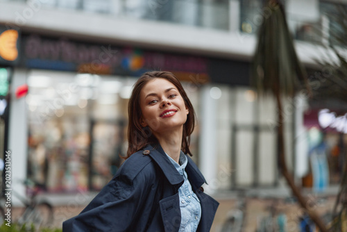 Photography Young woman with a bright smile stands outdoors on a city street, wearing a denim jacket and light shirt, candid urban portrait with blurred storefronts
