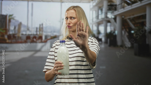 Foto Middle aged blonde woman holding milk bottle, palm outstretched in stop gesture inside bright airport terminal; boundary setting defiance