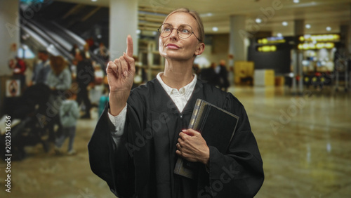 Quadro em tela Middle aged blonde woman judge in robe points finger while holding law book in busy airport terminal; authority resolve