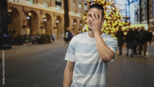 Fotografi Man covering face with hand while smiling in a decorated building with blurred christmas tree and shoppers; embarrassed amusement
