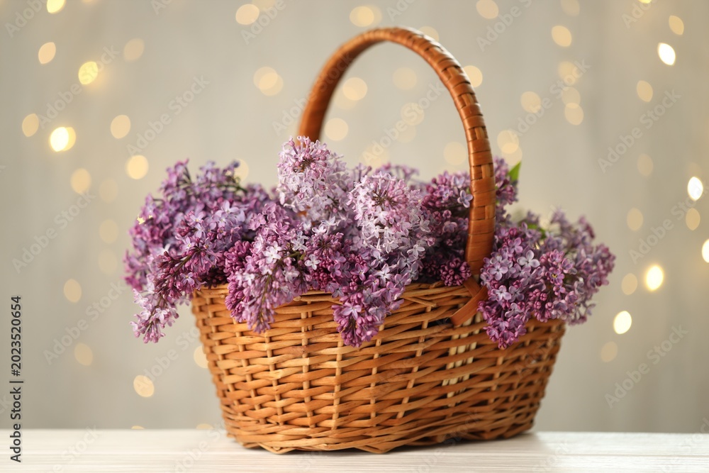 Fototapeta premium Fresh lilac flowers in wicker basket on white wooden table against grey background with blurred lights, closeup. Bokeh effect