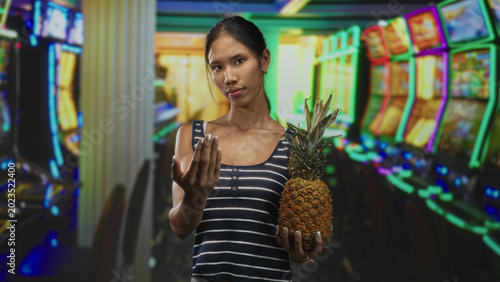 Photography Thai young woman in striped tank top points finger to camera while holding a pineapple among neon slot machines in a casino; defiance