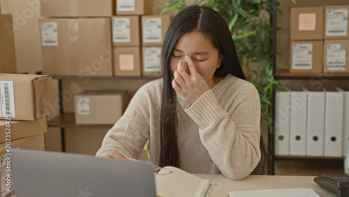 Foto Woman at a packing desk with laptop and cardboard parcels pinches nose and removes glasses while sorting orders in building; work stress