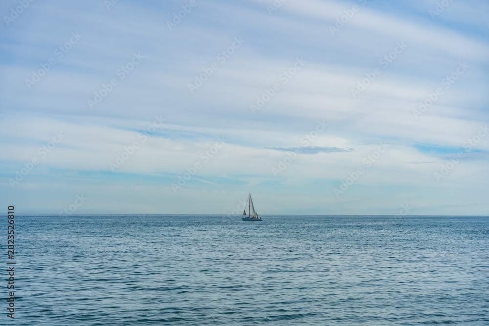Fototapeta premium Minimal seascape with small sailboat on calm blue ocean under soft cloudy sky. Peaceful marine travel and freedom concept.