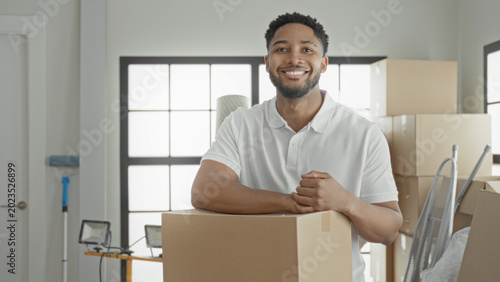 Obraz na plátně Smiling man holds a cardboard box, rests arm on it and raises fist beside stacked boxes and floodlight in building; moving day confidence