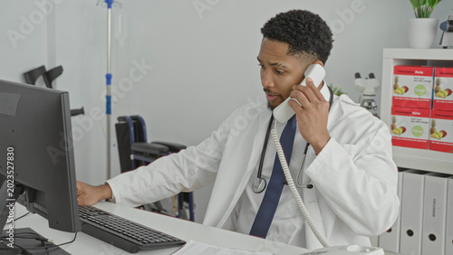 Fotografie Man holding phone to ear while typing on computer at a clinic desk, stethoscope visible; focused care