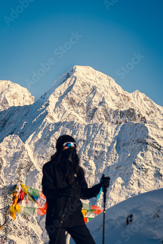 Blurred foreground of a young woman in black trekking attire standing on a mountain overlooking the Himalayas, Mardi Himal trekking, Nepal. The peaks are covered in snow, and the sky is clear.