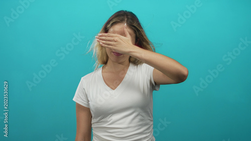 Canvas Print Woman covering her eyes with hand in studio against turquoise background, wearing white v neck t shirt, frontal pose; shy privacy