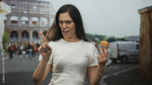 Photos Woman grimacing and finger pointing to cupcake on sunlit bustling street by ancient coliseum at midday; disgust