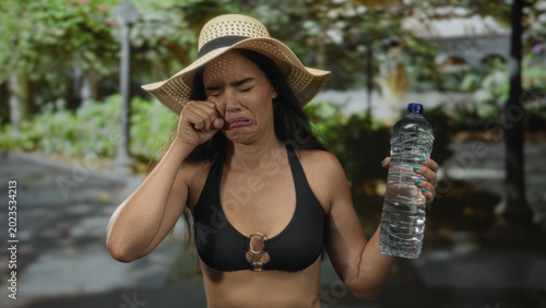 Canvas Print Woman wearing black bikini top and straw hat wipes tears with fist and holds water bottle in forest; sadness