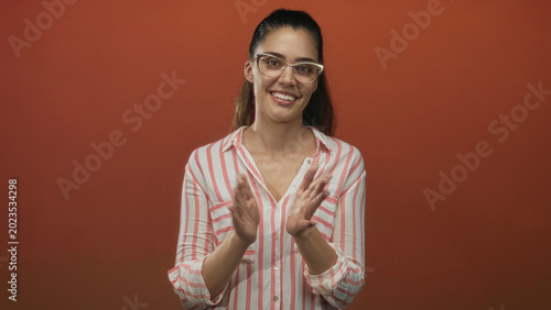 Obraz na plátně Woman smiling and clapping hands in orange studio, wearing glasses and a pink and white striped shirt with rolled sleeves; joy celebration