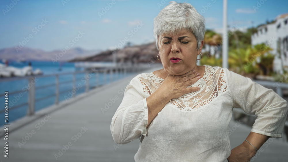 Fototapeta premium Woman clutching throat with hand on a seaside boardwalk pier by railing and boats, facial grimace and visible neck gesture showing breathing difficulty; distress.