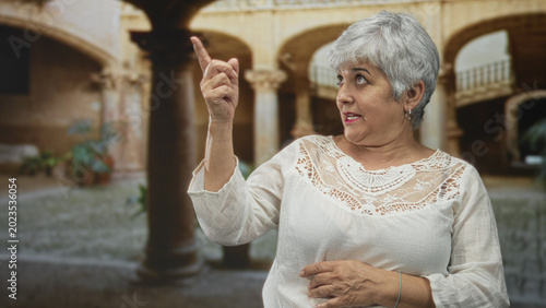 Slika na platnu Woman pointing finger, hand on chest, speaking, wearing white lace blouse and hoop earring in a historic building courtyard; curiosity memory