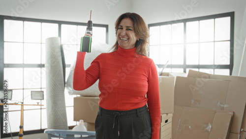Quadro su tela Woman holding power drill aloft while smiling among moving boxes and packing materials in a building; confidence