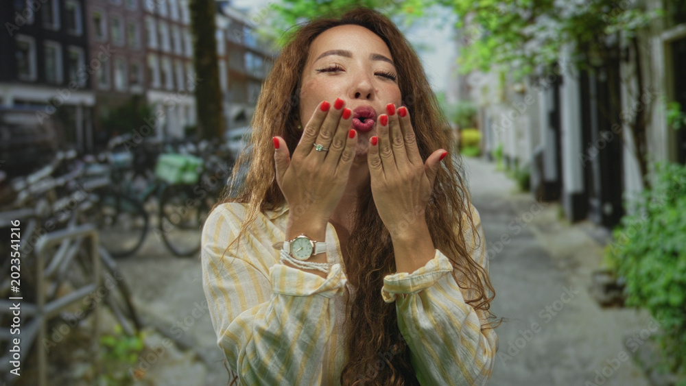 Fototapeta premium Woman blowing a kiss with both hands extended toward camera on a city street lined with bicycles and storefronts; playful affection.