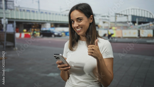 Foto Woman smiling while texting on smartphone on street, holding phone with both hands and briefly giving thumbs up; everyday happiness