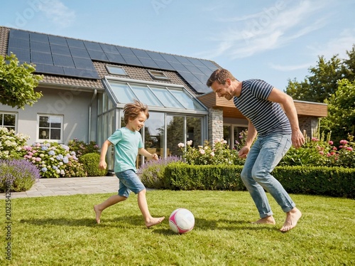 Father and son playing soccer barefoot on lush green lawn near modern house with solar panels
