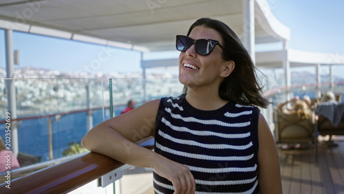 Bild auf Leinwand Young brunette woman lifts sunglasses and smiles, arm resting on wooden deck railing, hair blown by breeze, on ship building terrace with ocean view; serenity