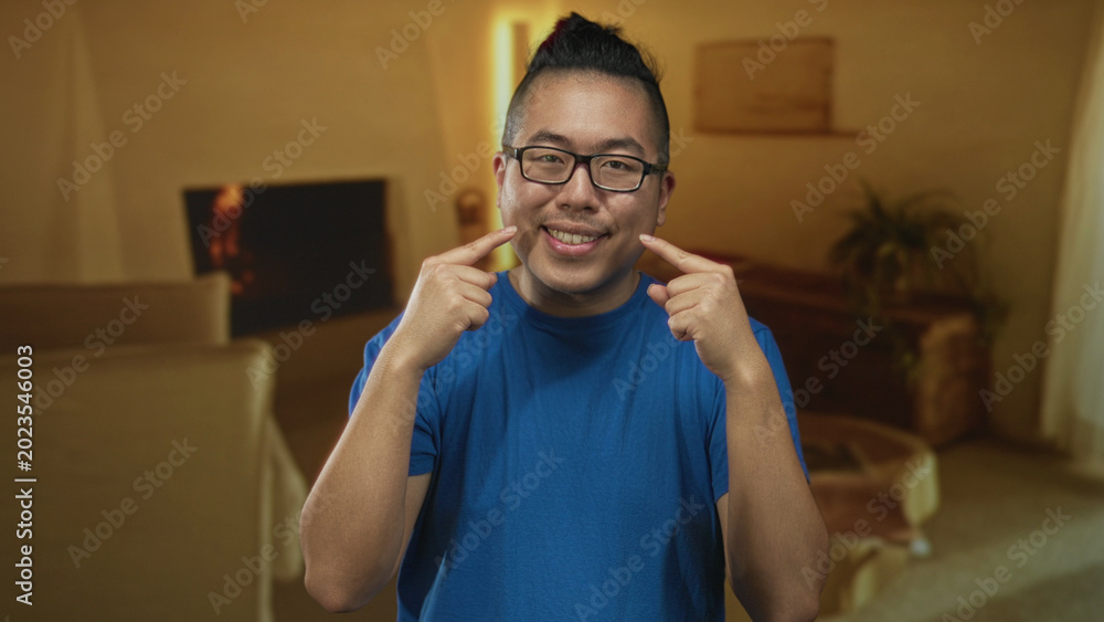 Obraz premium Man pointing fingers to cheeks in building living room scene showing smile and glasses; warm domestic happiness.