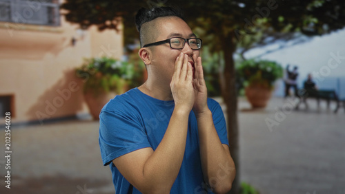 Foto Young man gesturing with hands and smiling on a street near a building, wearing a blue tshirt and glasses; joyful conversation