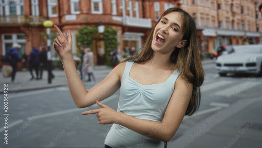 Fototapeta premium Young woman points finger on a city street crosswalk while smiling and gesturing with both hands; playful confidence.