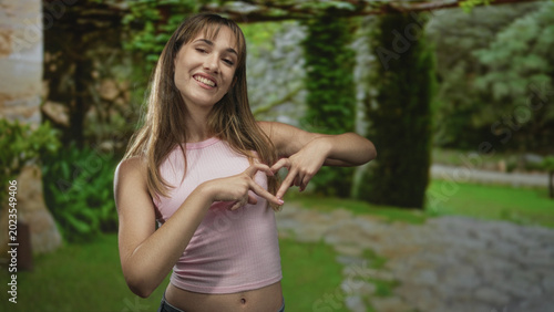Bild auf Leinwand Young woman in pink crop top makes heart shape with hands and shows navel in park with green trees and stone path; youthful joy warmth