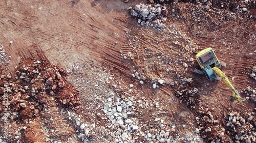 Aerial view of heavy construction machinery operating on rocky excavation site. Excavator moves across rough terrain scattered with stones and exposed earth
