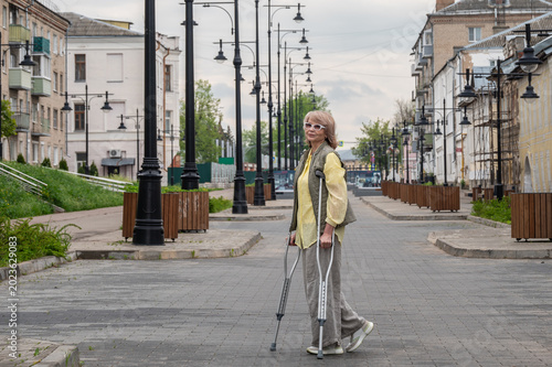 An adult attractive woman walks along a city street on crutches after an injury.