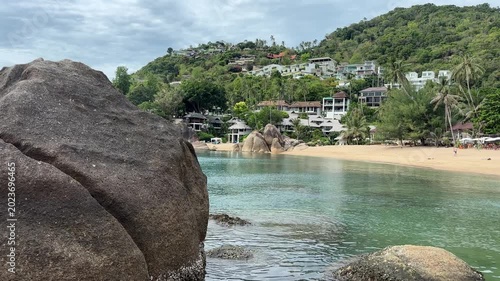 Turquoise bay with a sandy beach, large rocks, palm trees, and tropical hillside buildings, creating a peaceful island landscape and coastal travel destination in Thailand.