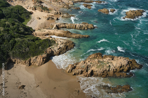 Aerial view of a rocky beach featuring golden sand and turquoise waves crashing on the shore amidst green vegetation.