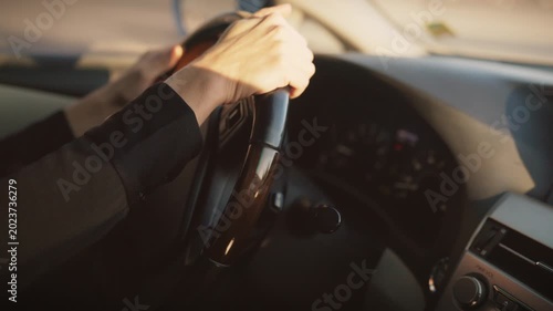 Close-up of driver’s hands holding steering wheel inside car during golden hour. Person wearing black sleeves controlling vehicle, dashboard and speedometer visible in soft sunlight, interior in focus