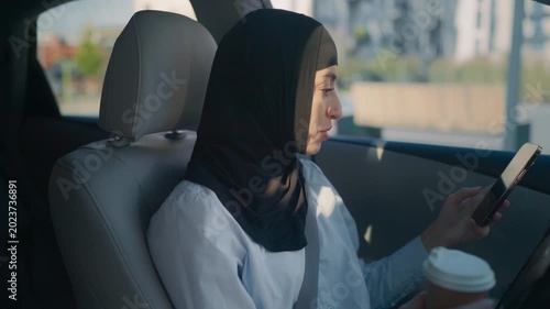 Middle Eastern beautiful woman wearing black hijab sitting in driver seat holding coffee cup and smartphone. Female driver inside modern car in daylight checking phone while parked or waiting.