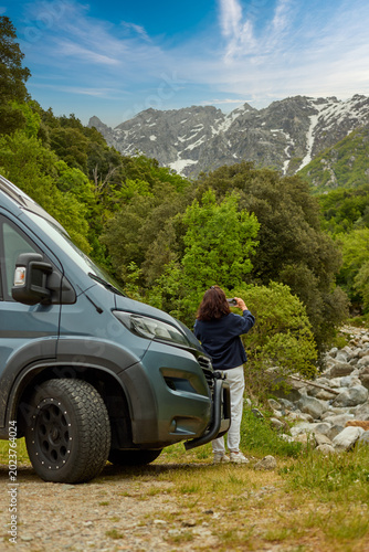 Active woman traveler standing beside a camper van and taking a photo of a mountain valley with forest and rocky peaks in Corsica. Colors: green, gray, blue.
