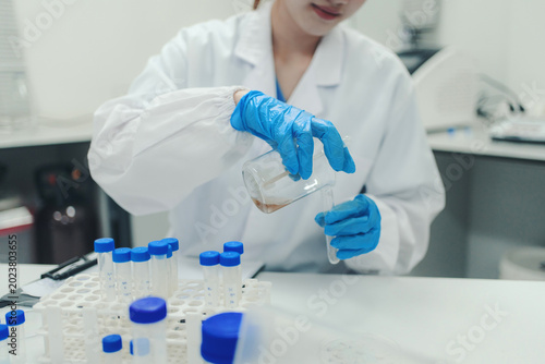 Scientist wearing a lab coat and blue gloves poring liquid from a beaker into a test tube in a laboratory environment