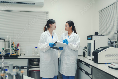 Two female scientists in lab coats and safety glasses discussing data on a tablet in a modern laboratory setting