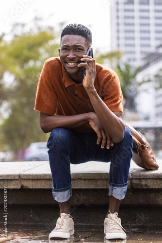 Man crouching on stone ledge by water at plaza, holding phone, rust polo, backpack beside