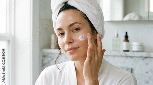 Woman gently applies a white moisturizing cream to her left cheek with her finger while looking forward in a clean, bright bathroom after showering and wrapping her hair in a white towel